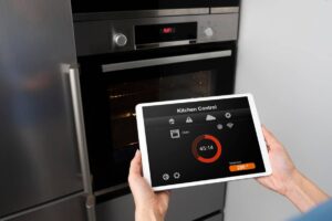 Hands holding a tablet showing a kitchen control app, with a modern oven in the background.