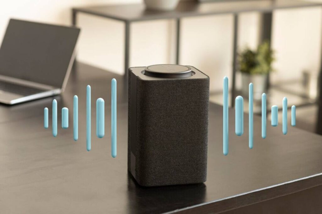 Smart speaker on a desk with visual sound waves, beside a laptop in a modern room.