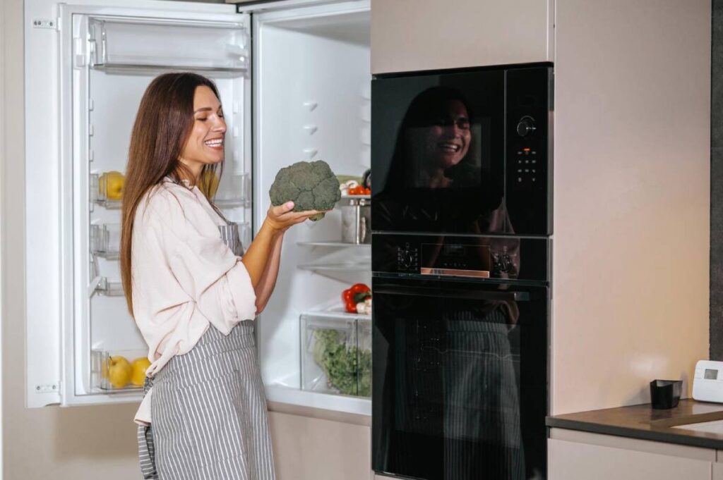 Woman in apron smiling at a broccoli beside an open fridge in a modern kitchen.