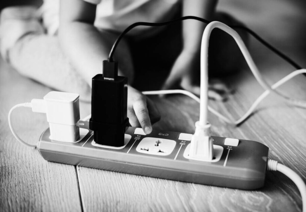 A child plugs various cables into a surge protector on the floor. Black and white photo.