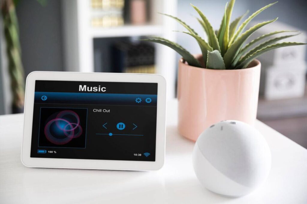 White tablet displaying a music app with a "Chill Out" playlist, beside a pink potted aloe vera and a white spherical speaker.