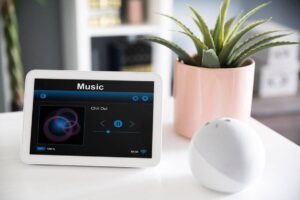 White tablet displaying a music app with a "Chill Out" playlist, beside a pink potted aloe vera and a white spherical speaker.