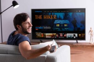 Man smiling and holding a remote, sitting on a sofa facing a TV screen displaying a movie menu titled "THE HIKER".