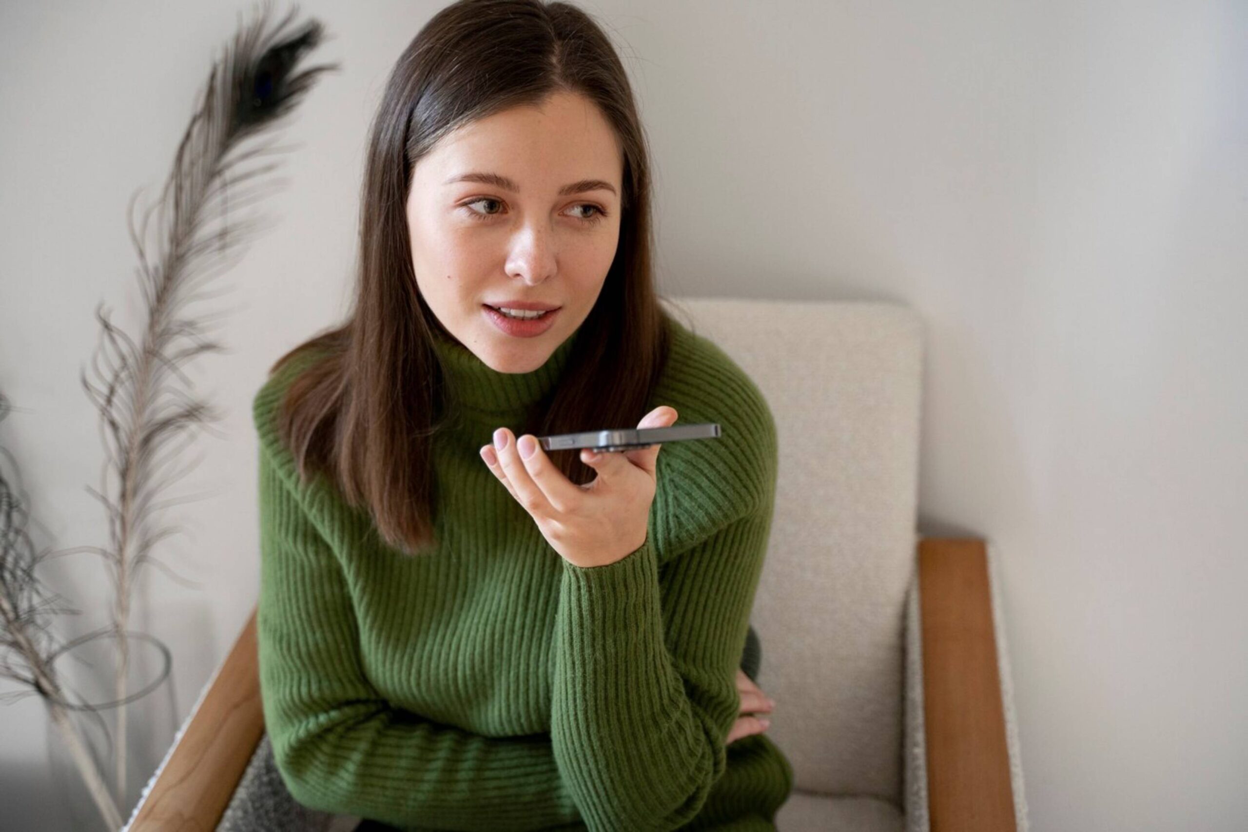 Young woman in green turtleneck speaking into smartphone, sitting in a light, modern room. 