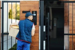 Delivery man in a blue uniform entering a code on a digital keypad by a modern door, holding a parcel.