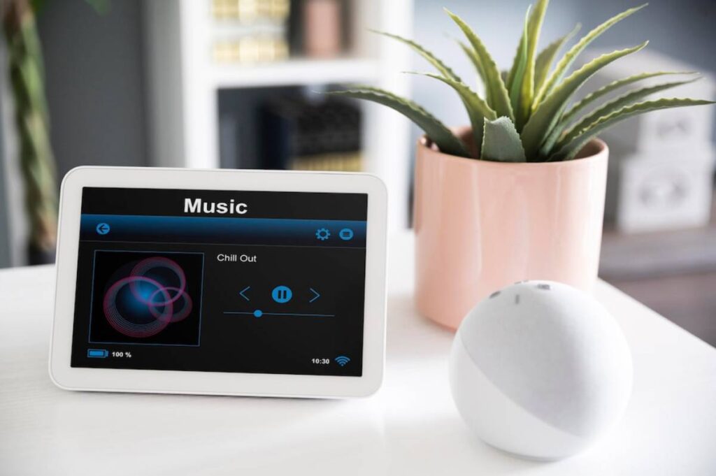 Smart display showing music interface on a table next to a potted plant and a smart speaker.