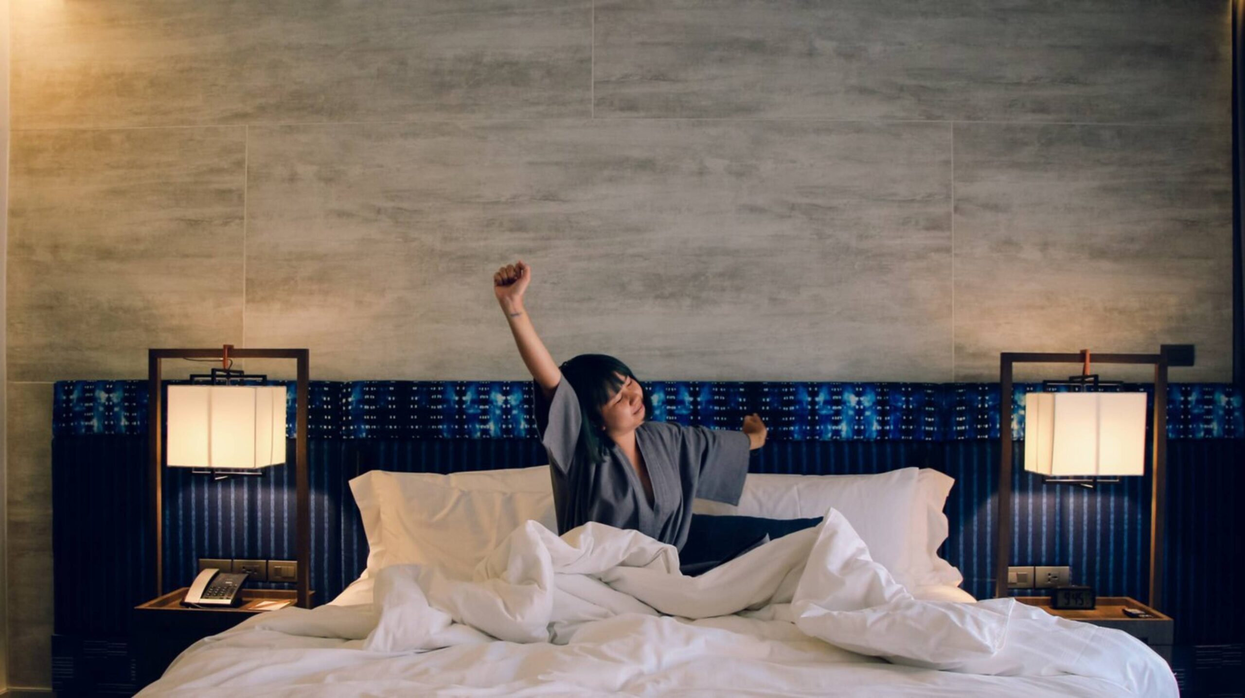 A woman stretches joyfully in a messy hotel bed with a serene grey wall backdrop and bedside lamps.