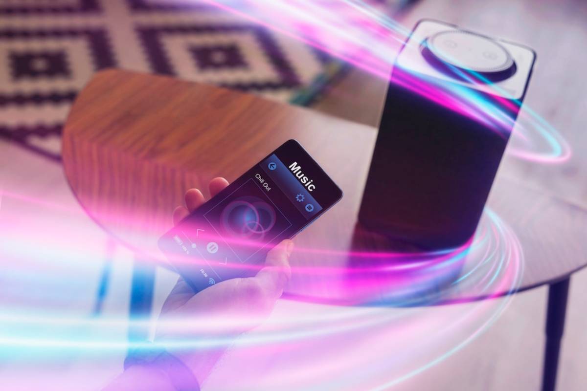 A hand holding a smartphone controlling a wireless speaker with colorful light waves, indicating music playback in a modern setting. 