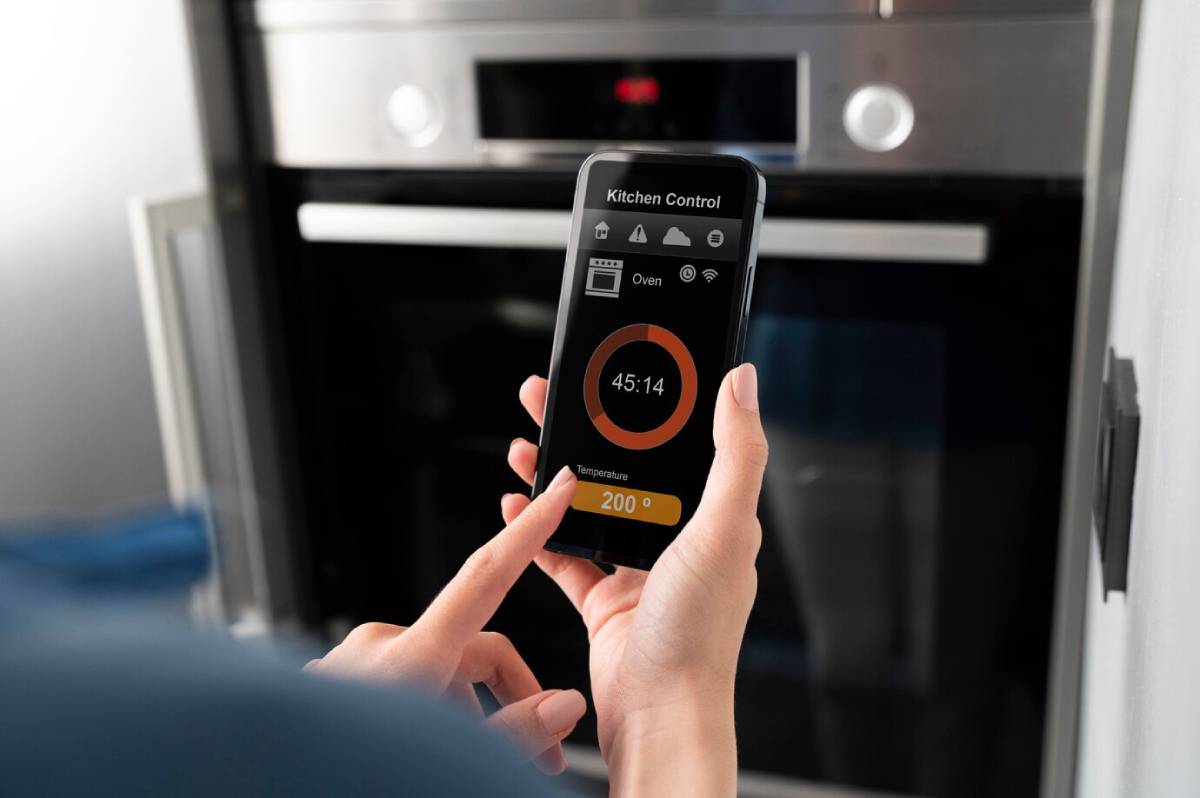 Hands using a smartphone to control an oven remotely with a digital interface displaying temperature and timer. 