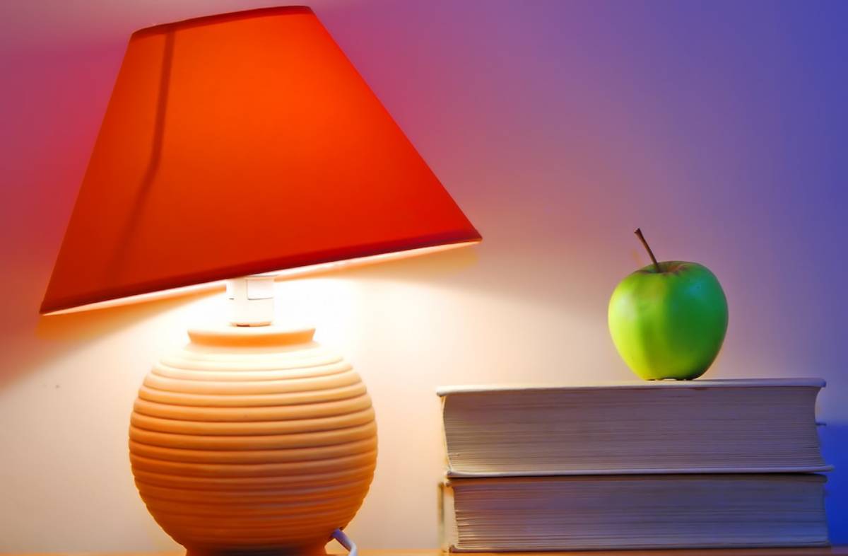A table lamp with a red shade illuminates a stack of books with a green apple on top.