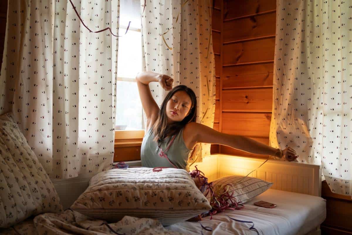 Young woman yawning and stretching in bed, in a cosy, sunlit wooden cabin room.