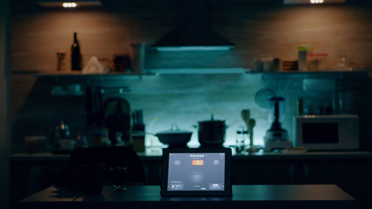 A dimly lit modern kitchen with an illuminated smart home control tablet on the counter. 