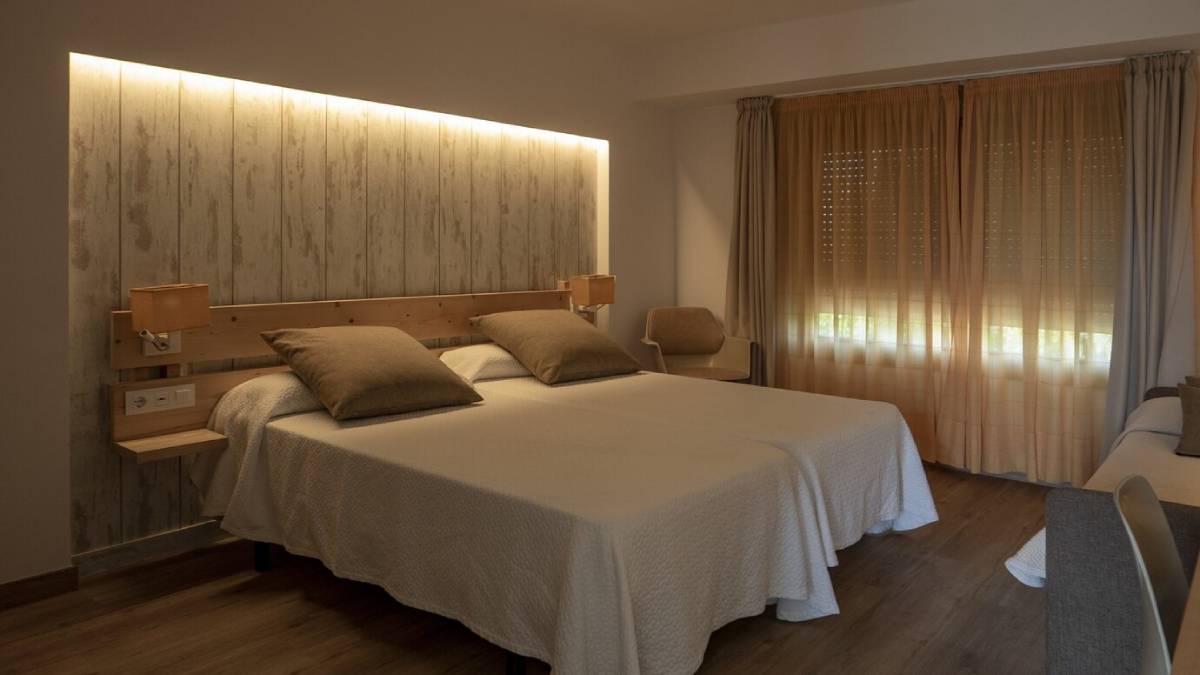 A cosy bedroom with a large bed, wooden headboard, subdued lighting, and sheer curtains filtering sunlight. 