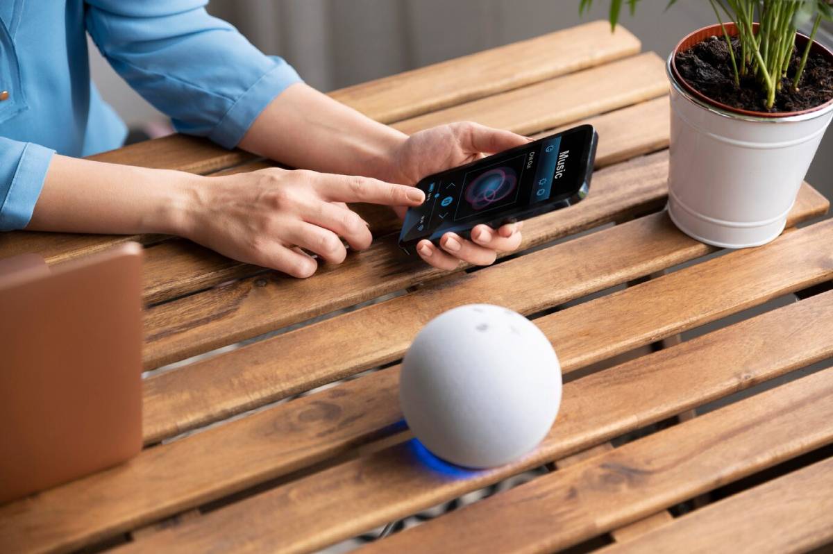 Person using smartphone to control a smart speaker on a wooden table, with a potted plant nearby. 