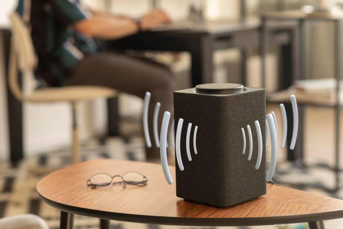  A smart speaker on a table, visually emitting sound waves, with eyeglasses nearby and a person in the background.