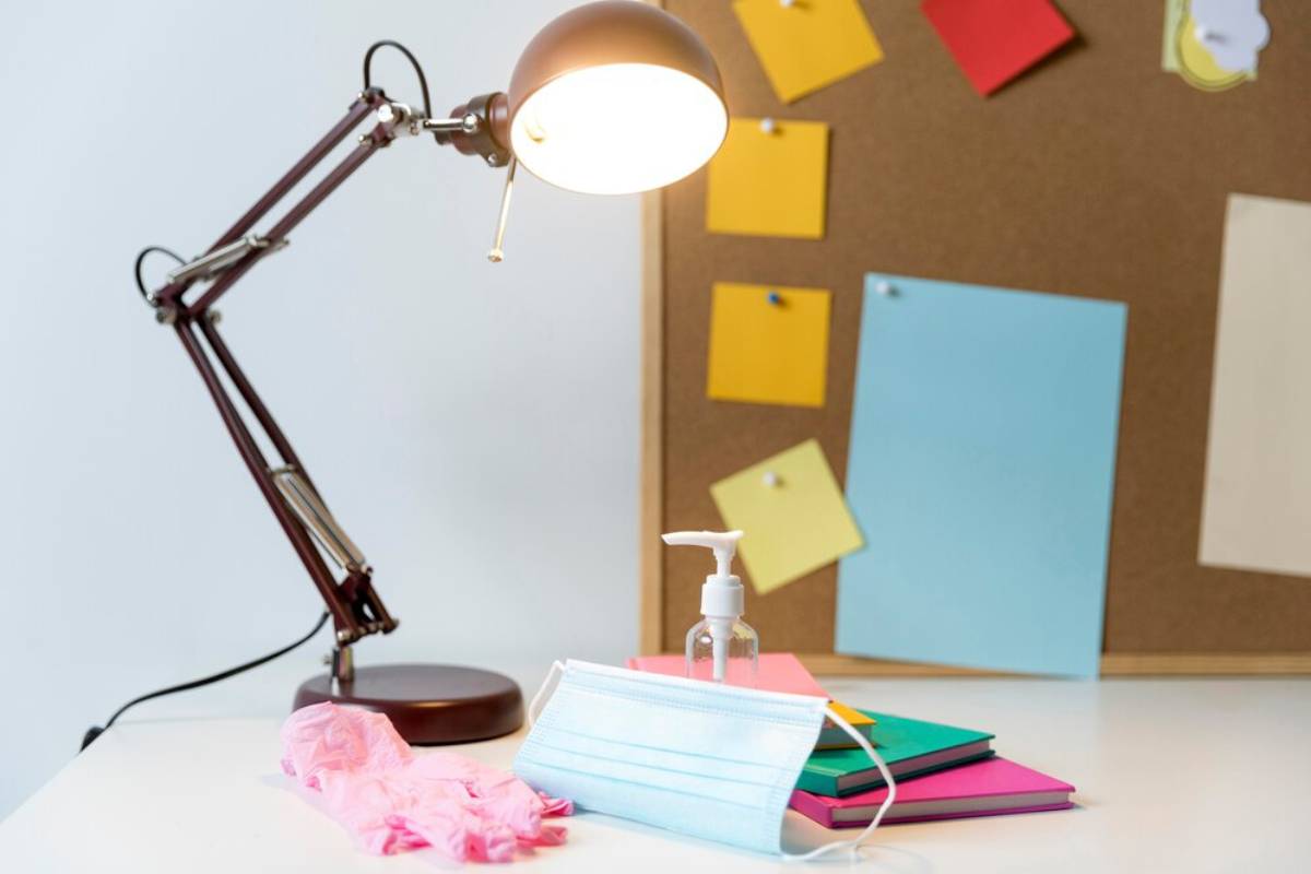 A workspace featuring a desk lamp, stacked notebooks, a face mask, hand sanitizer, and a pinboard with colourful notes.