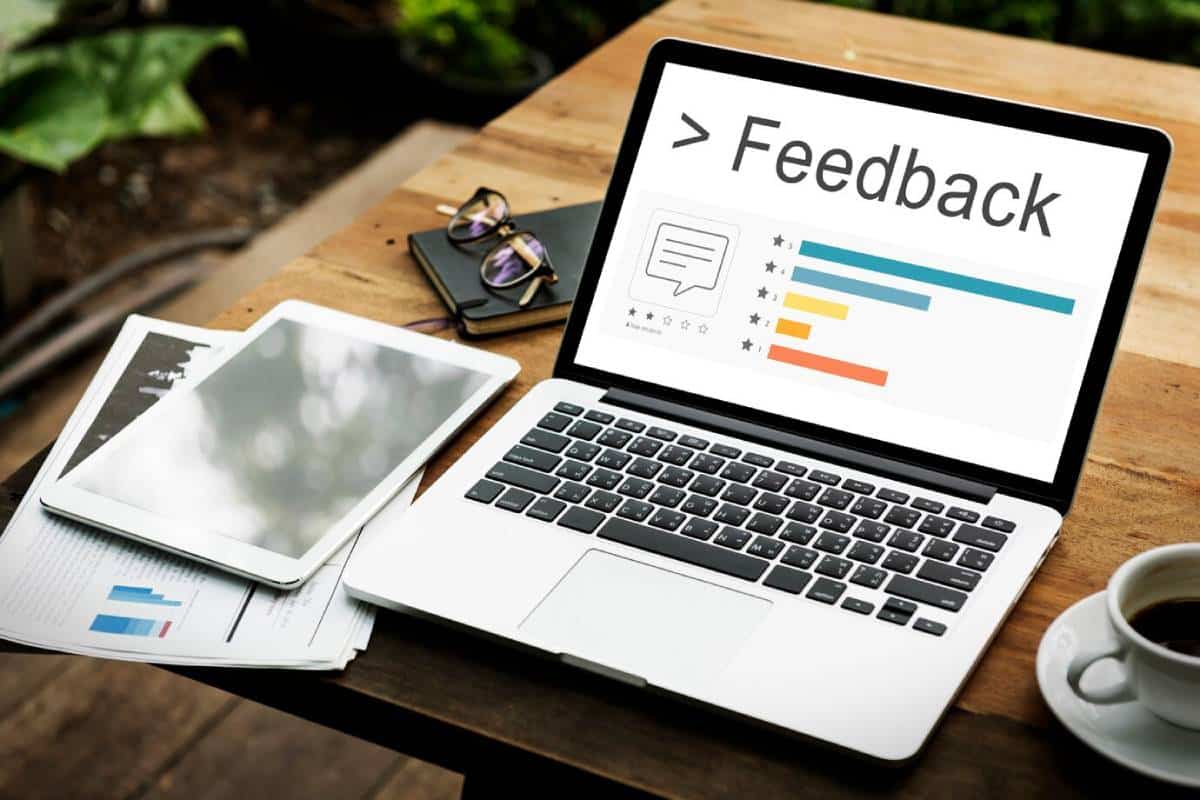 Laptop displaying a feedback chart on a wooden table with documents, sunglasses, and a cup of coffee.
