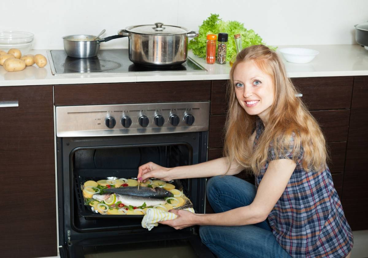 Woman in kitchen placing a tray with a seasoned fish into the oven, smiling at the camera.