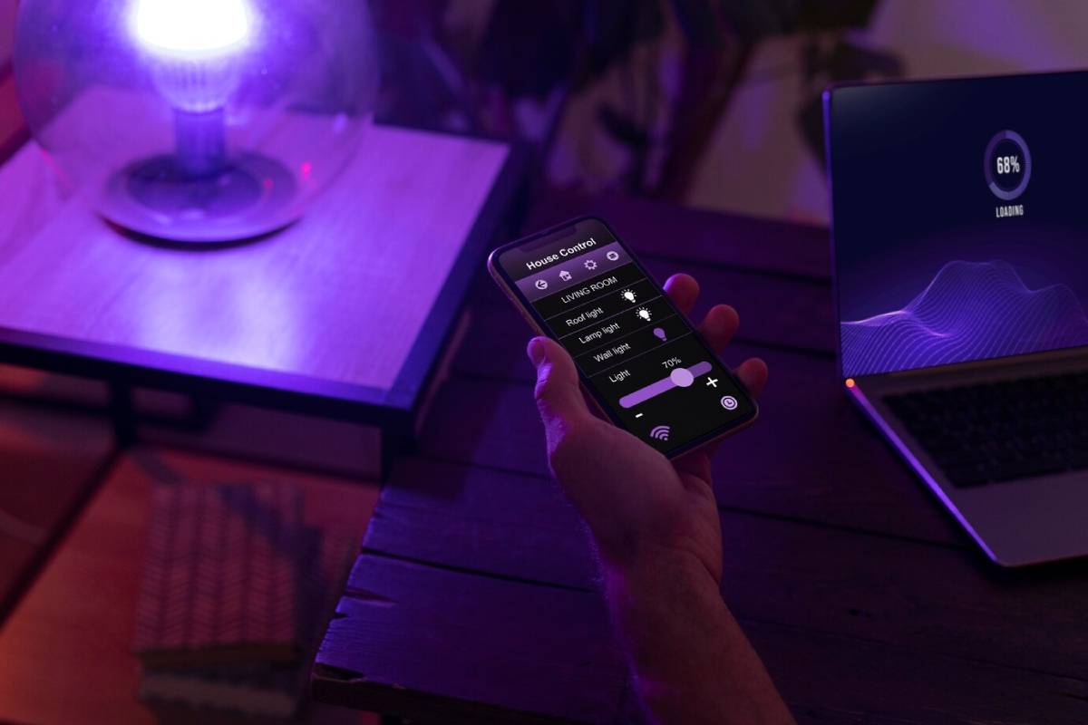 Person using a smartphone to control home devices like lights, visible in a dimly lit room with a laptop in the background. 