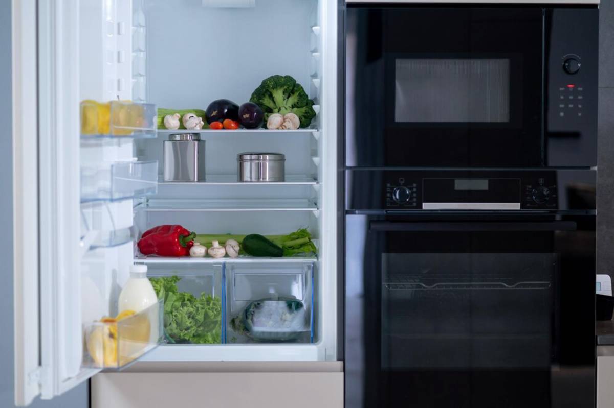 Open fridge displaying vegetables and other food items next to a built-in microwave and oven.