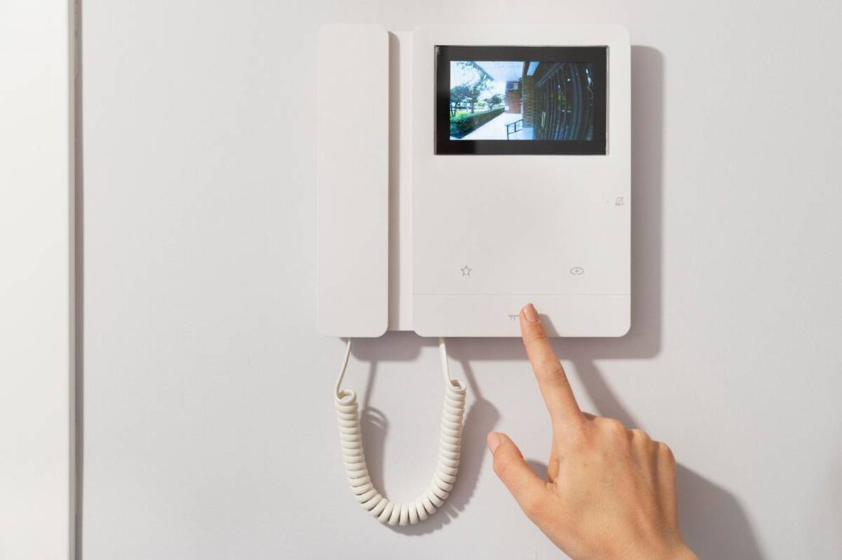 A person's hand pressing a button on a white video intercom displaying an outdoor scene. 