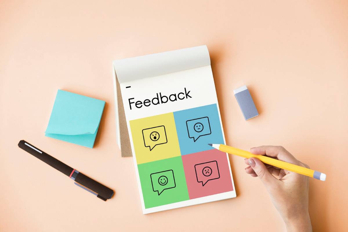  Hand holding a pencil over a feedback form with colorful emoji icons on a light pink background.