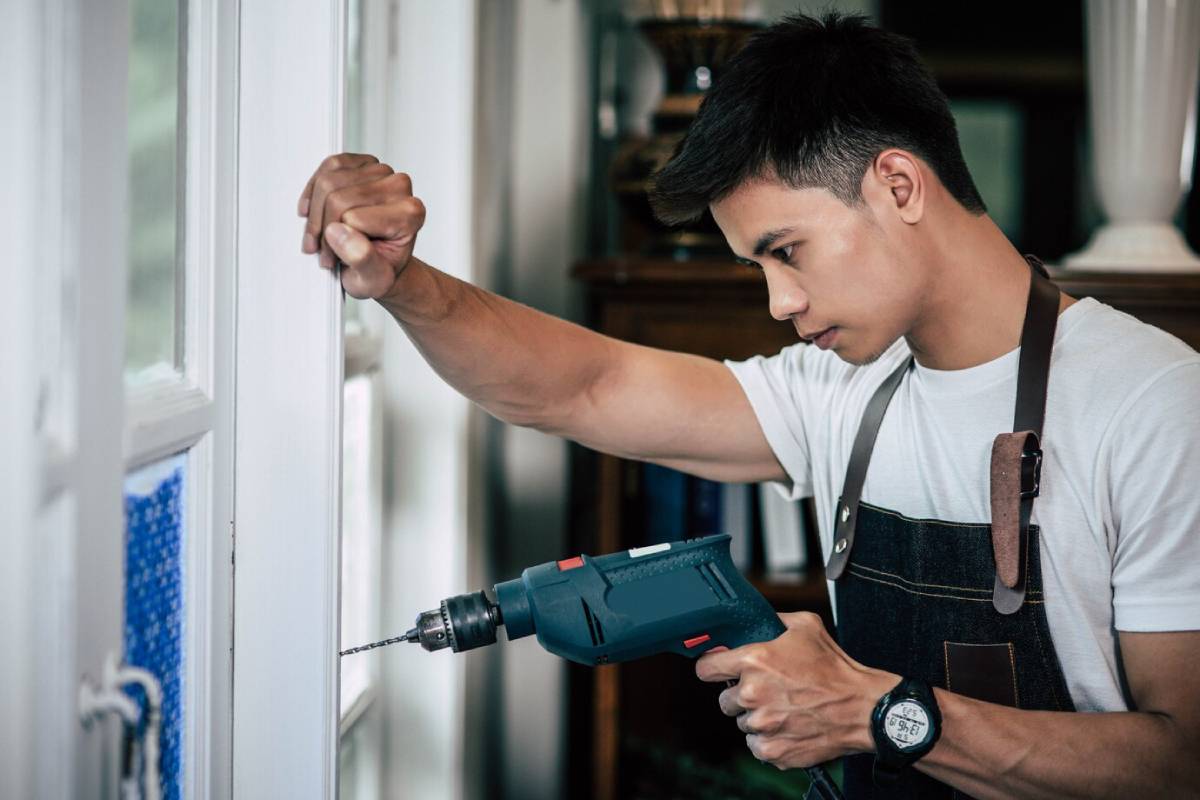  Young man drilling into a door frame, wearing a white t-shirt and brown tool belt.