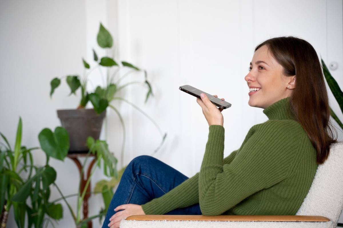 A woman in a green sweater smiling while talking on her smartphone, indoor setting with houseplants.