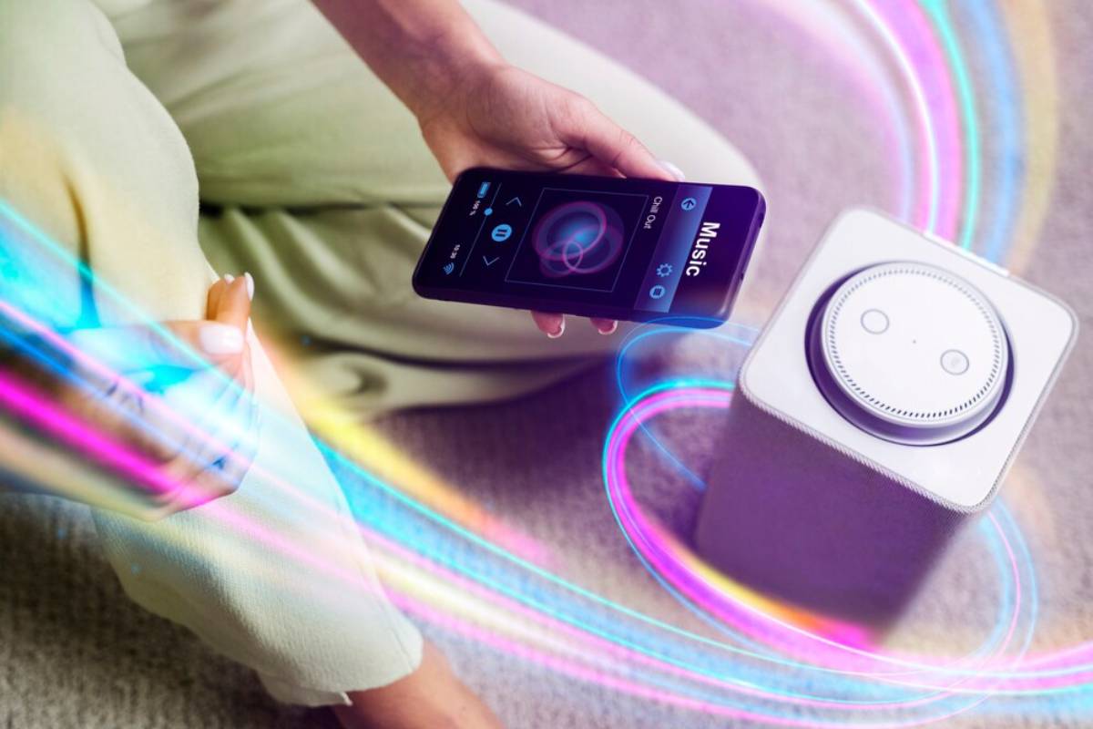 Person using a smartphone to control a smart speaker with colourful sound waves emanating from both devices.