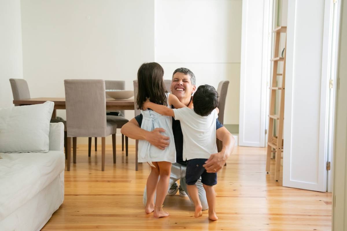A man joyfully embracing two young children in a bright living room. 