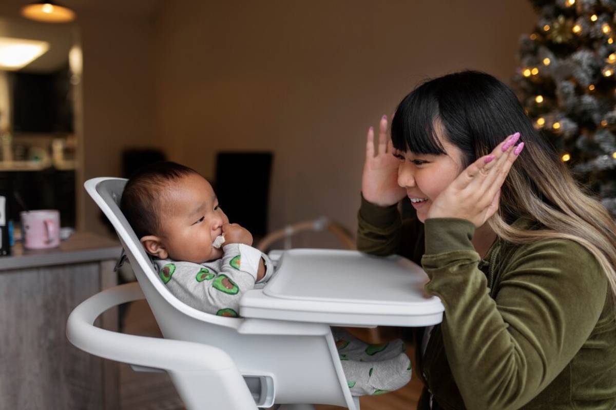 Woman playing peekaboo with a baby in a high chair, with a Christmas tree in the background. 