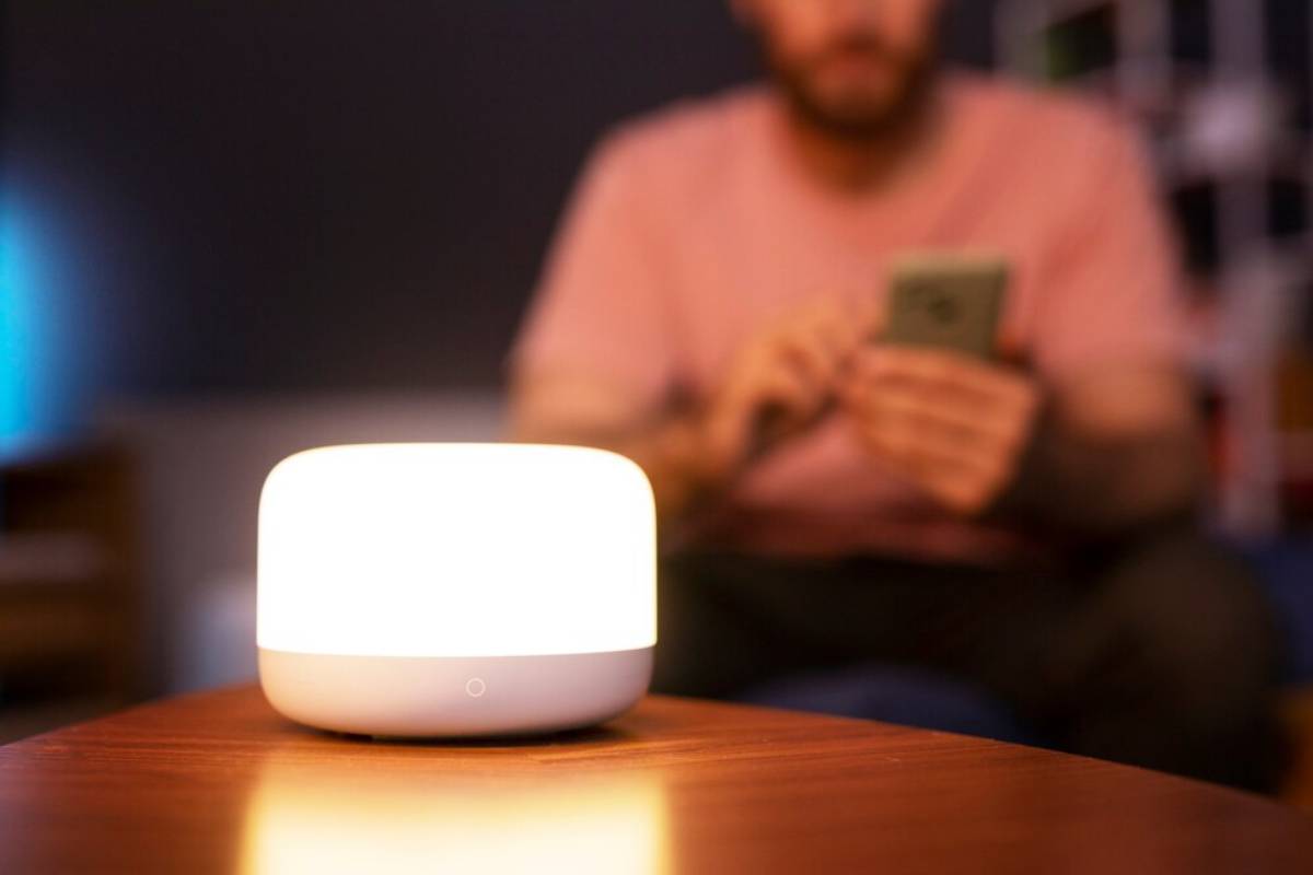 Illuminated smart home device on table, with blurred man using smartphone in background. 