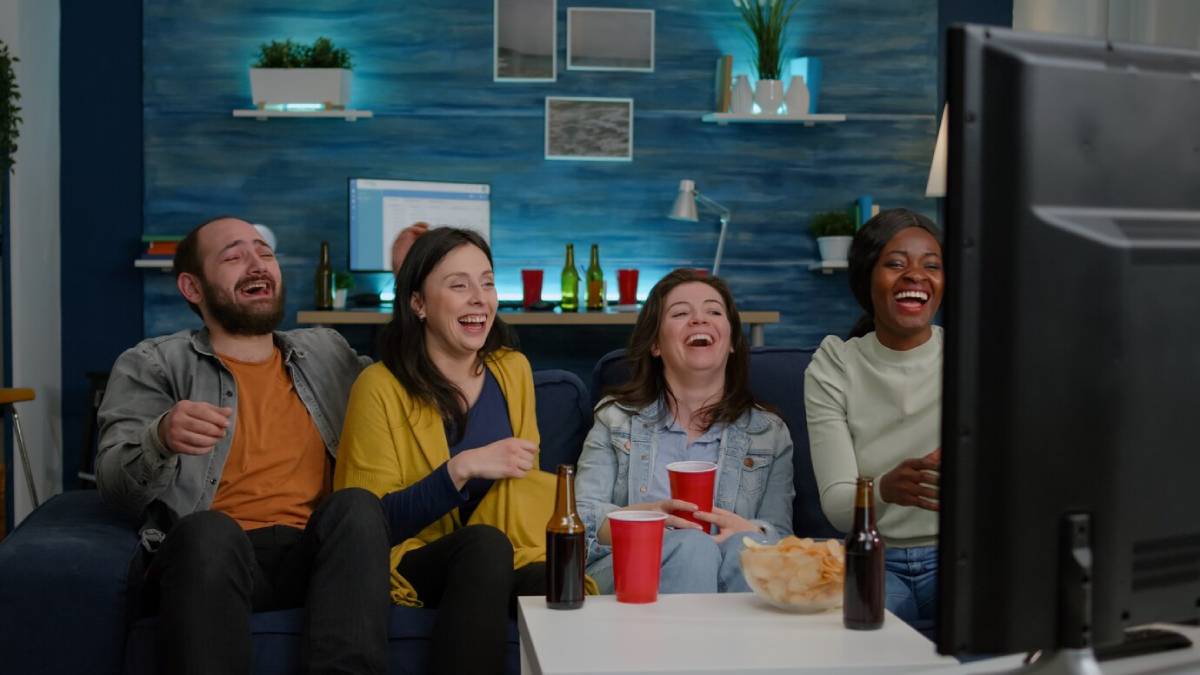 Four friends laughing while watching TV in a cozy living room with blue walls and modern decor. 