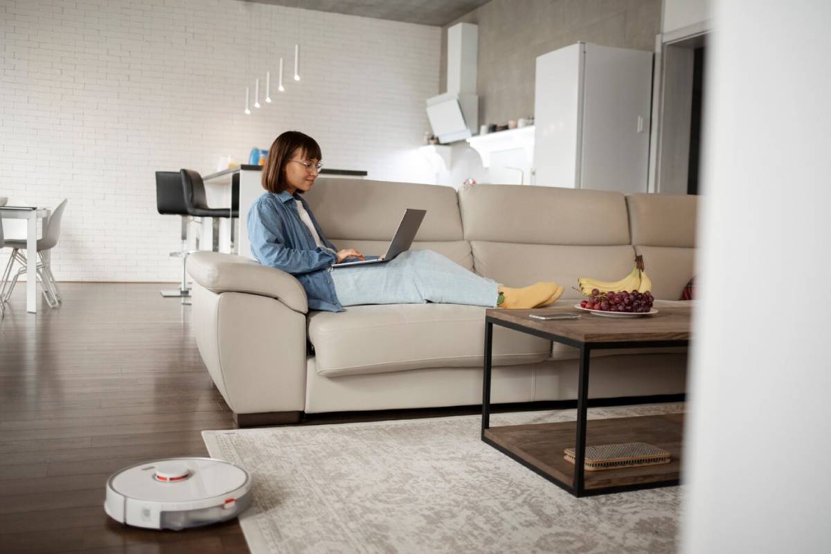 A woman working on a laptop on a sofa, in a modern living room, with a robotic vacuum cleaner active on the floor.