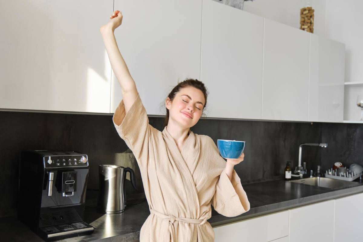Woman in beige bathrobe stretches joyfully while holding a blue mug in a modern kitchen.