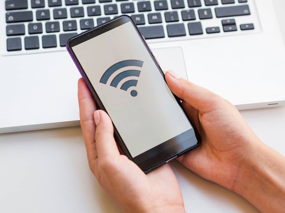  Person holding a smartphone displaying a Wi-Fi signal icon, with a laptop in the background.