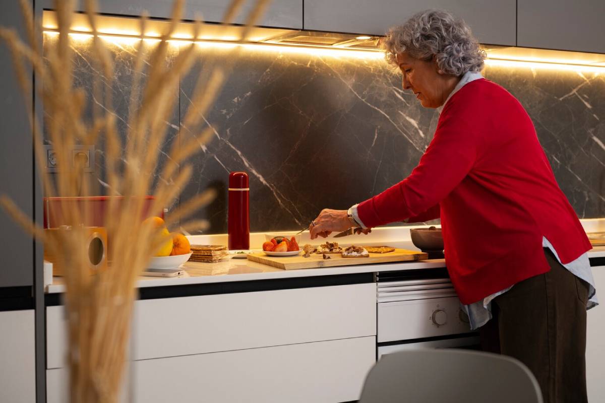 Elderly woman in a red cardigan slicing fruit in a modern kitchen.